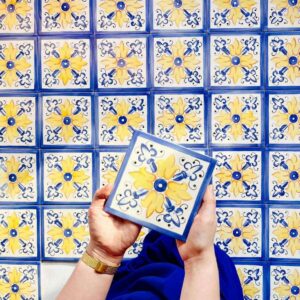 Żółto-niebieski nasycony ornament namalowany ręcznie na kafelkach ceramicznych, jedna sztuja trzymana w rękach autorki